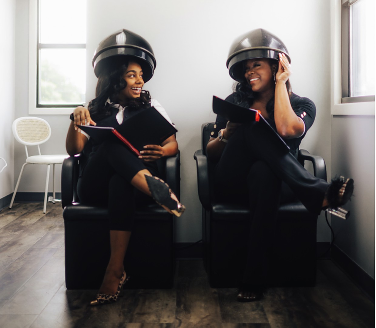 Two guests seated under hooded salon dryers, holding folders and smiling at each other